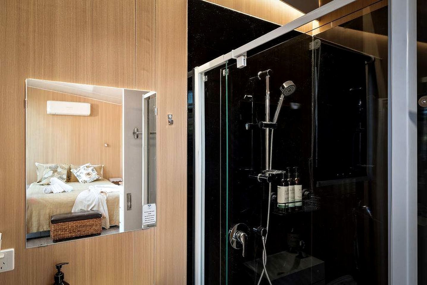 A modern bathroom with a glass shower enclosure, a wooden vanity, and a mirror reflecting a bed with pillows.