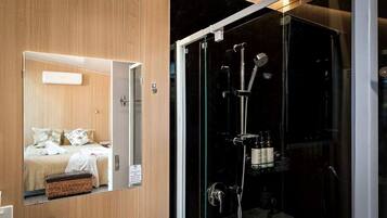 A modern bathroom with a glass shower enclosure, a wooden vanity, and a mirror reflecting a bed with pillows.