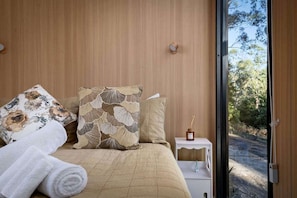A bedroom with a bed, pillows, a bedside table with a bottle, and a view of the outdoors.