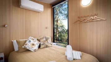 A bedroom with a bed, pillows, a bedside table, an air conditioning unit, a window with a view of trees, and a wall-mounted light fixture.