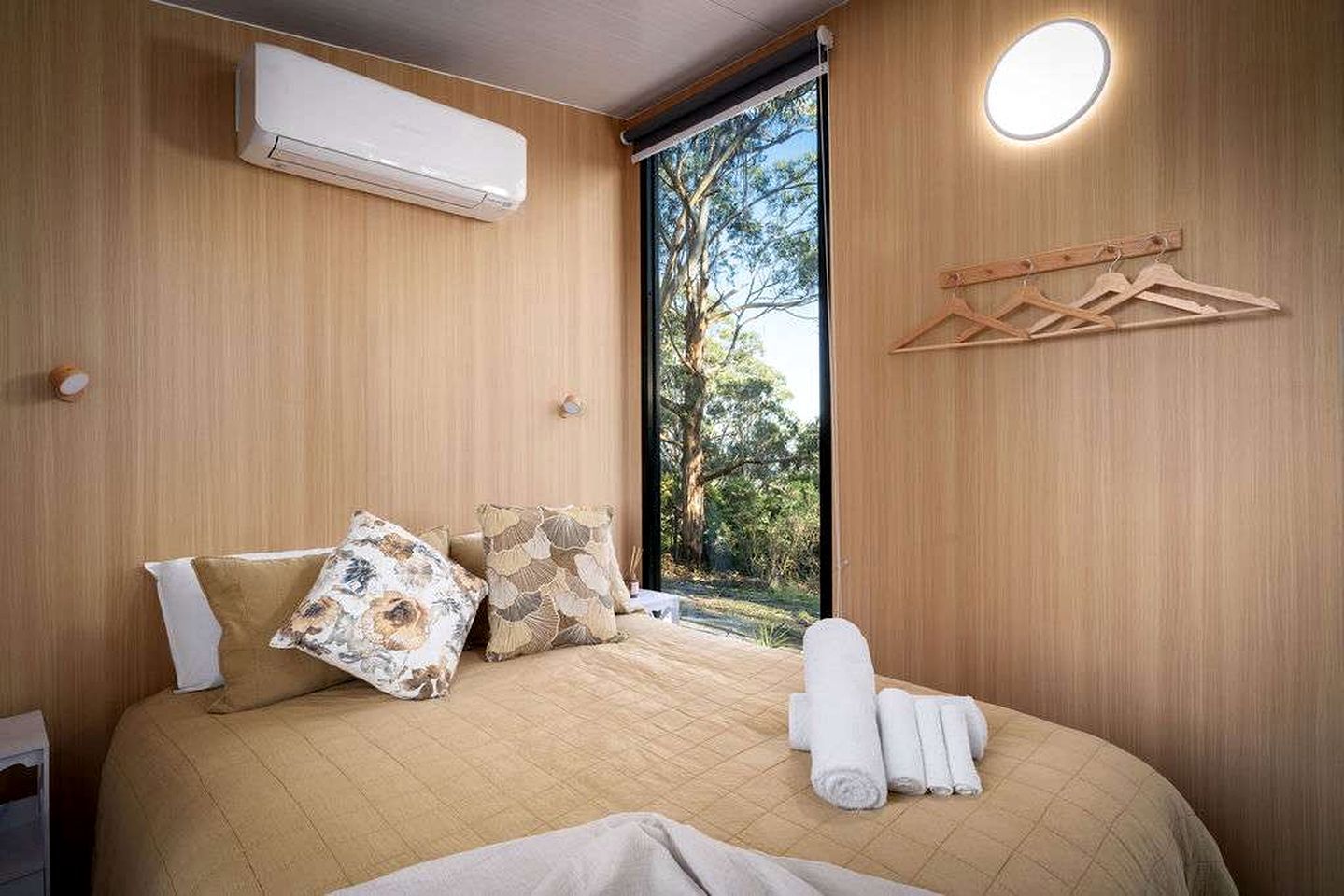 Une chambre à coucher comprenant un lit, des oreillers, une table de chevet, un appareil de climatisation, une fenêtre donnant sur des arbres et un luminaire fixé au mur.