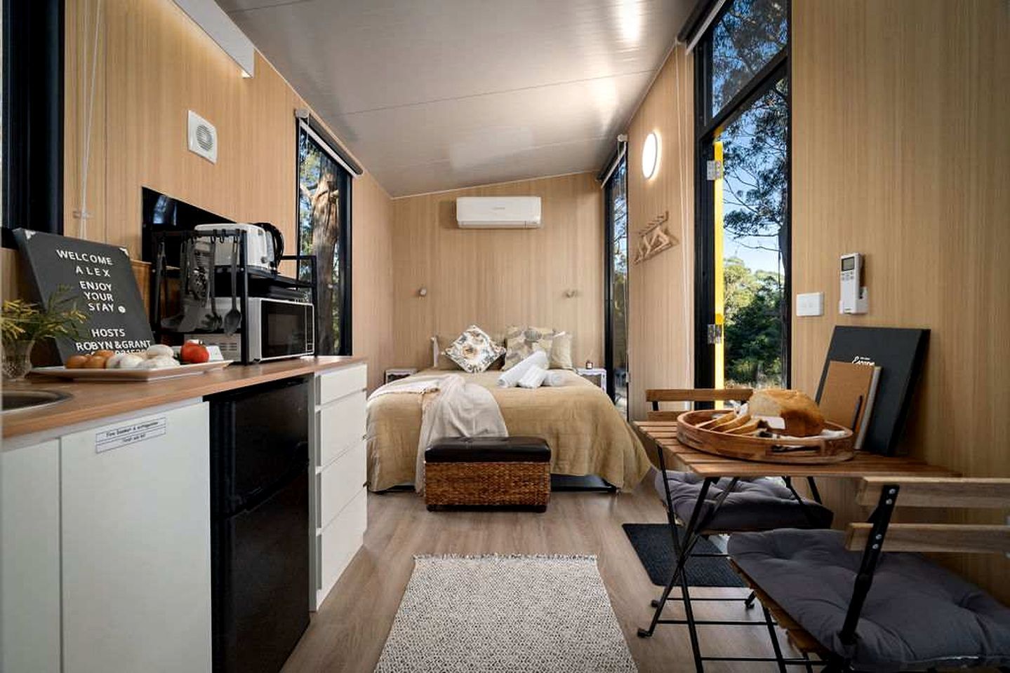 A compact hotel room with a kitchenette, a bed with a headboard, a dining area with a table and chairs, and a large window offering a view of the outdoors.
