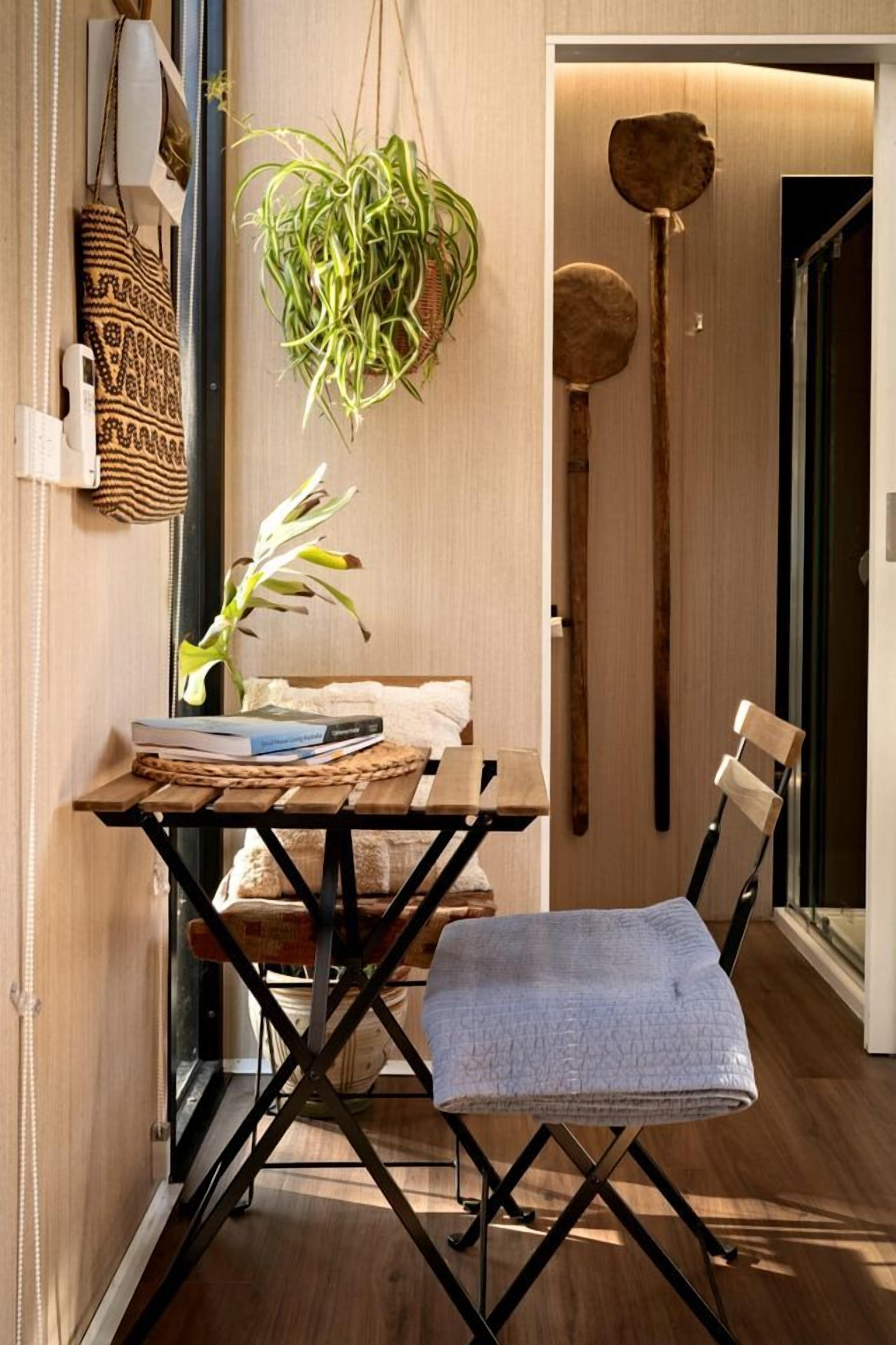 A compact room with a wooden floor, a folding table with a plant, a chair, and a wall-mounted shelf with a woven bag.