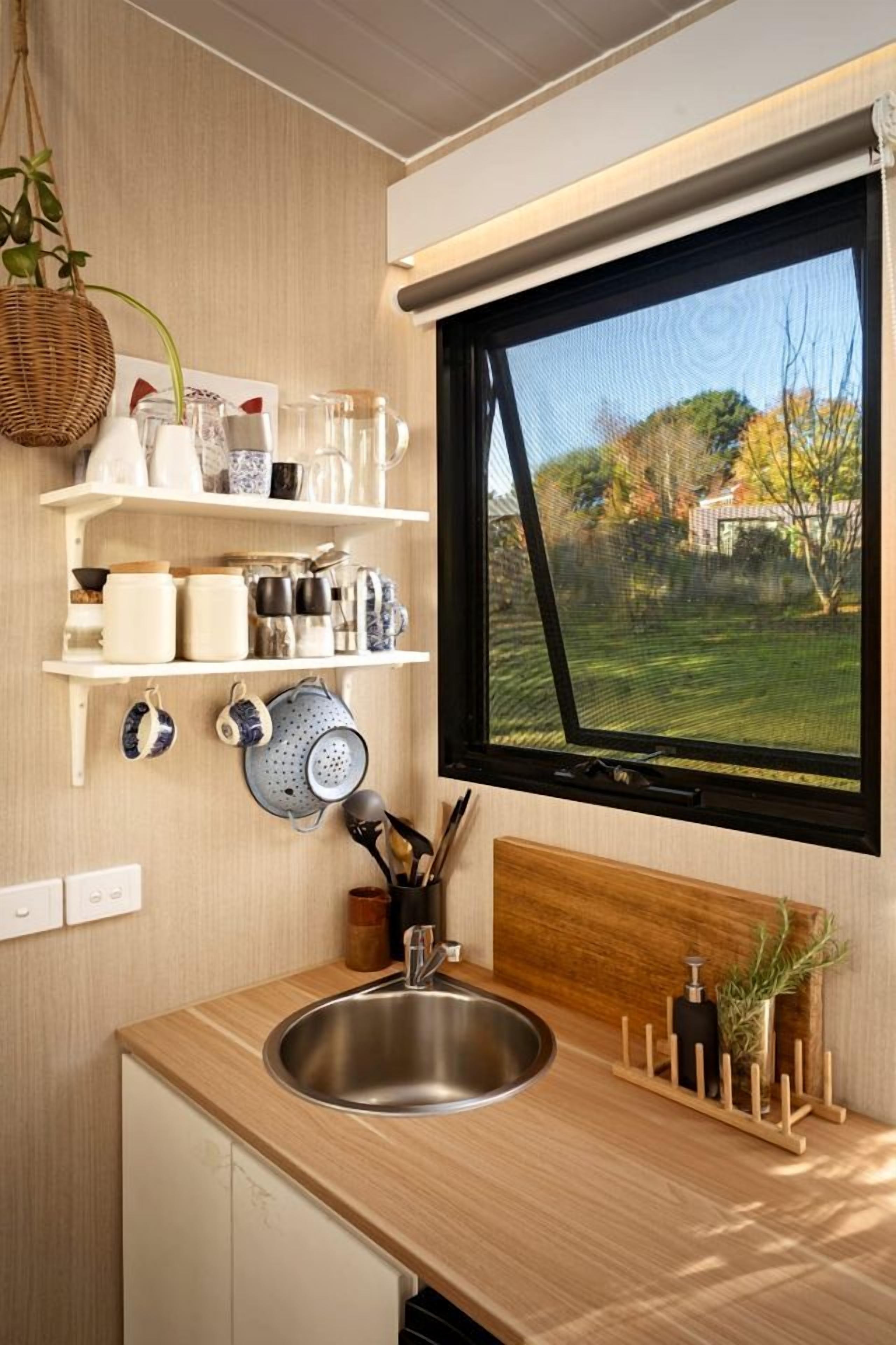 A kitchen with a stainless steel sink, wooden countertop, hanging shelves with kitchenware, and a window with a view of a garden.