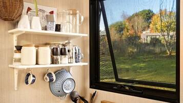 A kitchen with a stainless steel sink, wooden countertop, hanging shelves with kitchenware, and a window with a view of a garden.