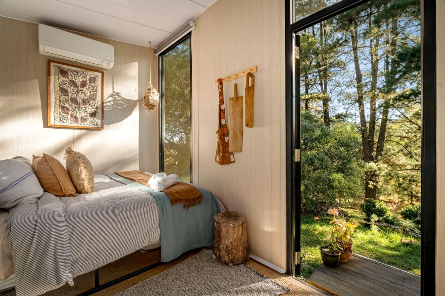 Une chambre avec une grande fenêtre, un lit avec des oreillers, une corbeille suspendue et un tabouret en bois.