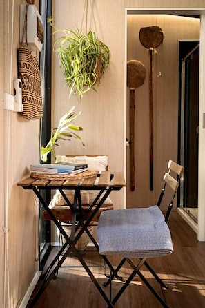Habitación pequeña con piso de madera, una mesa plegable con una planta, una silla y un estante en la pared con un bolso tejido.