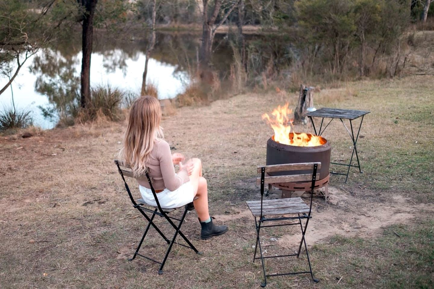 Une personne assise sur une chaise pliante près d’un feu de camp, dans un environnement naturel, avec une étendue d’eau en arrière-plan.