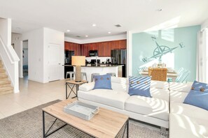 A modern living room with a sofa, coffee table, and a kitchen in the background. - Waterfront Family Mansion w BBQ, Games Room & Pool (Kissimmee)
