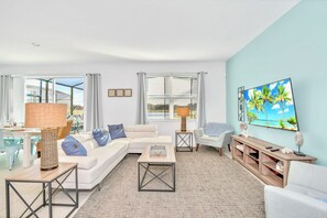 Un salon moderne avec un canapé blanc, une table basse, un téléviseur et une grande fenêtre équipée de stores.
