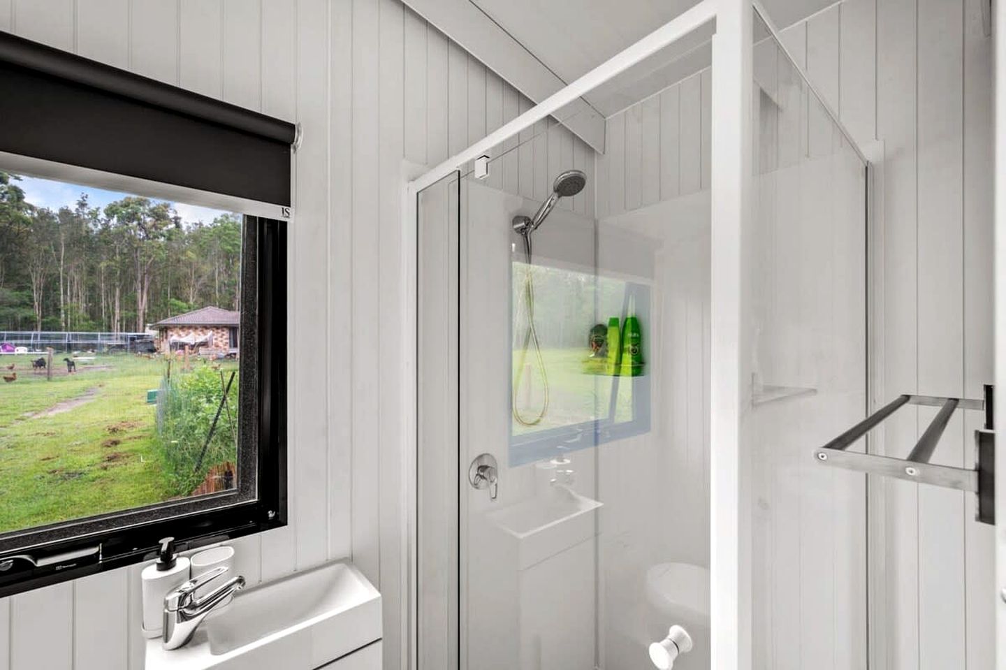 Un baño moderno con cabina de ducha de vidrio, un lavabo con grifo y una ventana que ofrece vistas a un área con césped y árboles.