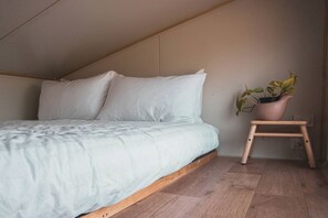 Et soveværelse med skrå væg, en seng med hvidt sengetøj, trægulv og en lille skammel med en potteplante.