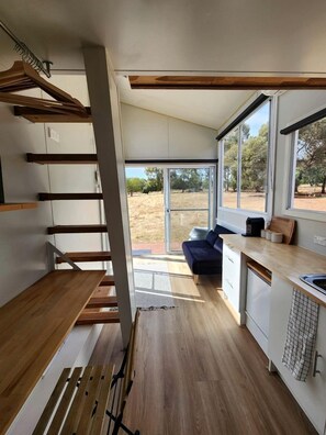 A modern kitchen with wooden flooring, a built-in bench, and large windows offering a view of the outdoors. - Charming Tiny House Overlooking the Table Top Mountains and Surrounded by Nature in Jindera, New South Wales (Jindera)