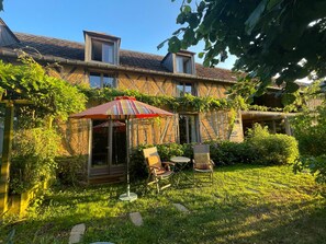 Une maison avec un jardin, une terrasse avec des chaises et un parasol à rayures.