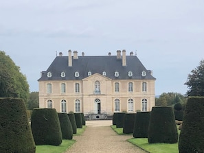 Un grand bâtiment symétrique, doté d’une entrée centrale et entouré de haies soigneusement taillées.
