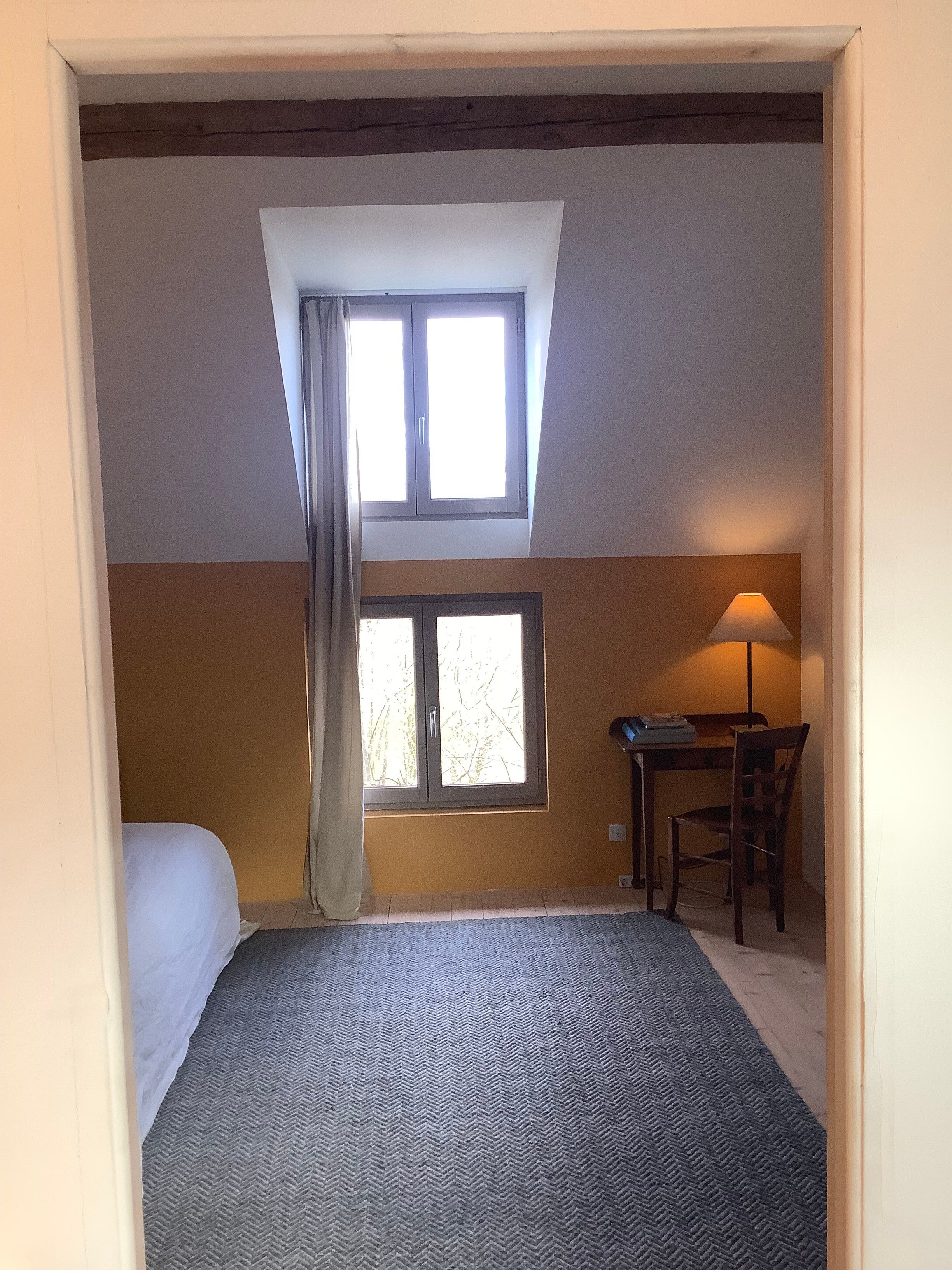 A bedroom with a bed, a desk, and a chair. There is a window with curtains and a lamp on the desk.