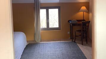 A bedroom with a bed, a desk, and a chair. There is a window with curtains and a lamp on the desk.