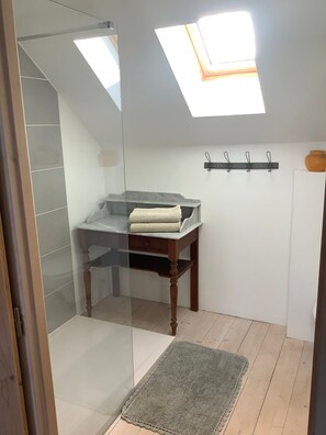 Un baño con tragaluz, cabina de ducha de vidrio, una mesa de madera con una toalla y un panel de pared con ganchos.
