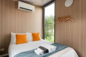A hotel room with a bed, an air conditioning unit, a window with a view of greenery, and a wooden hanger rack.