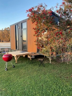 A modern outdoor seating area with a wooden table and chairs, a barbecue grill, and a small deck. - Tiny House Stay with Cozy Fireplace and Nature Views in Koonoomoo, Australia (Koonoomoo)