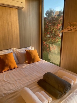 A hotel room with a large bed, pillows, towels, a window with a view of trees, and a wooden ceiling. - Tiny House Stay with Cozy Fireplace and Nature Views in Koonoomoo, Australia (Koonoomoo)
