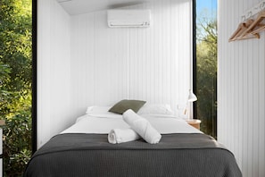 A bedroom with a bed, pillows, a blanket, a towel, an air conditioning unit, a nightstand, and a window with a view of greenery.