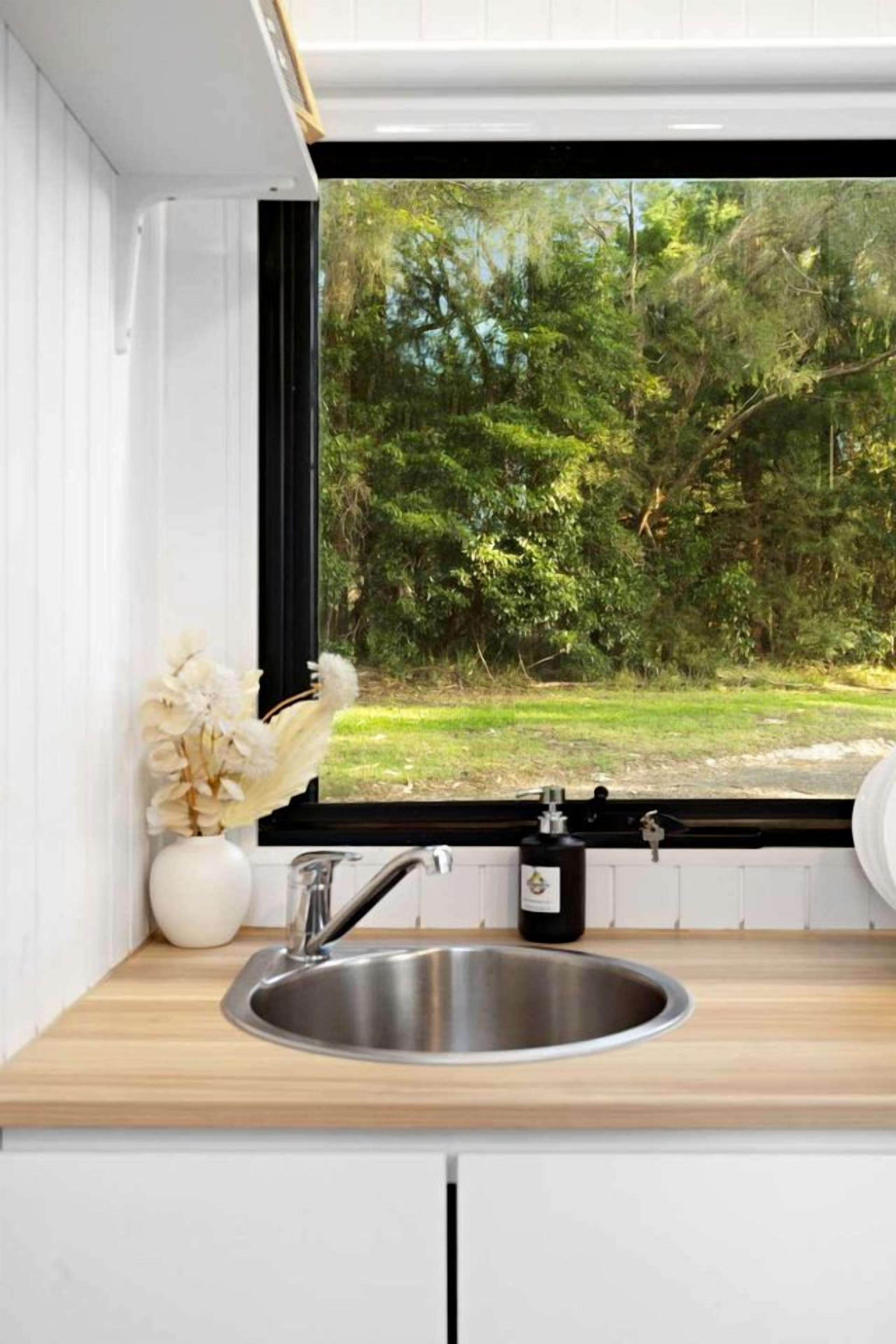 A kitchen with a stainless steel sink, a white vase with flowers, and a window overlooking a green landscape.