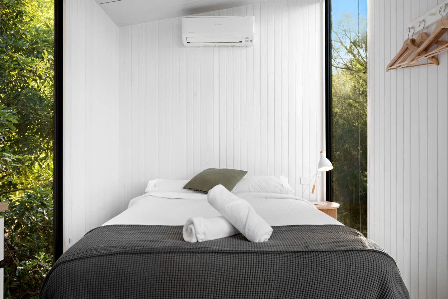 Un dormitorio con cama, almohadas, manta, toalla, equipo de aire acondicionado, mesita de noche y ventana con vista a vegetación.