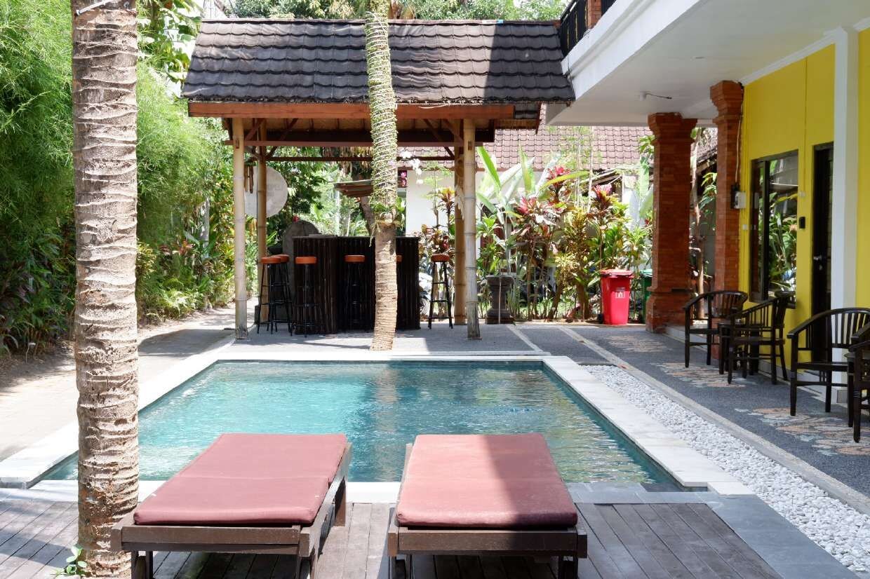 A pool area with two lounge chairs, a covered seating area, and a yellow building in the background.