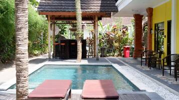 A pool area with two lounge chairs, a covered seating area, and a yellow building in the background.