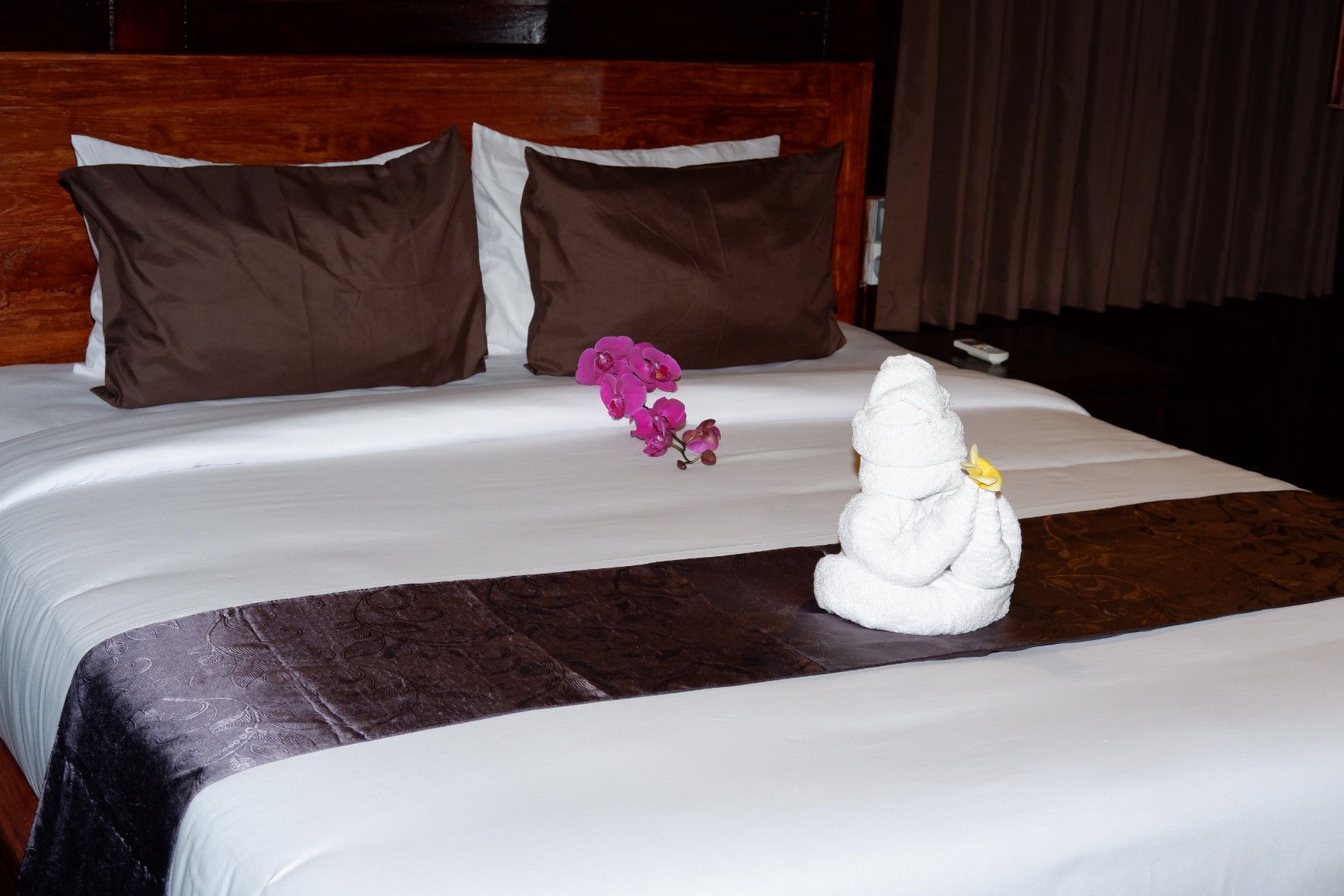 Un lit bien fait, avec une couette blanche, deux oreillers marron, une sculpture décorative en forme de serviette et des fleurs roses.