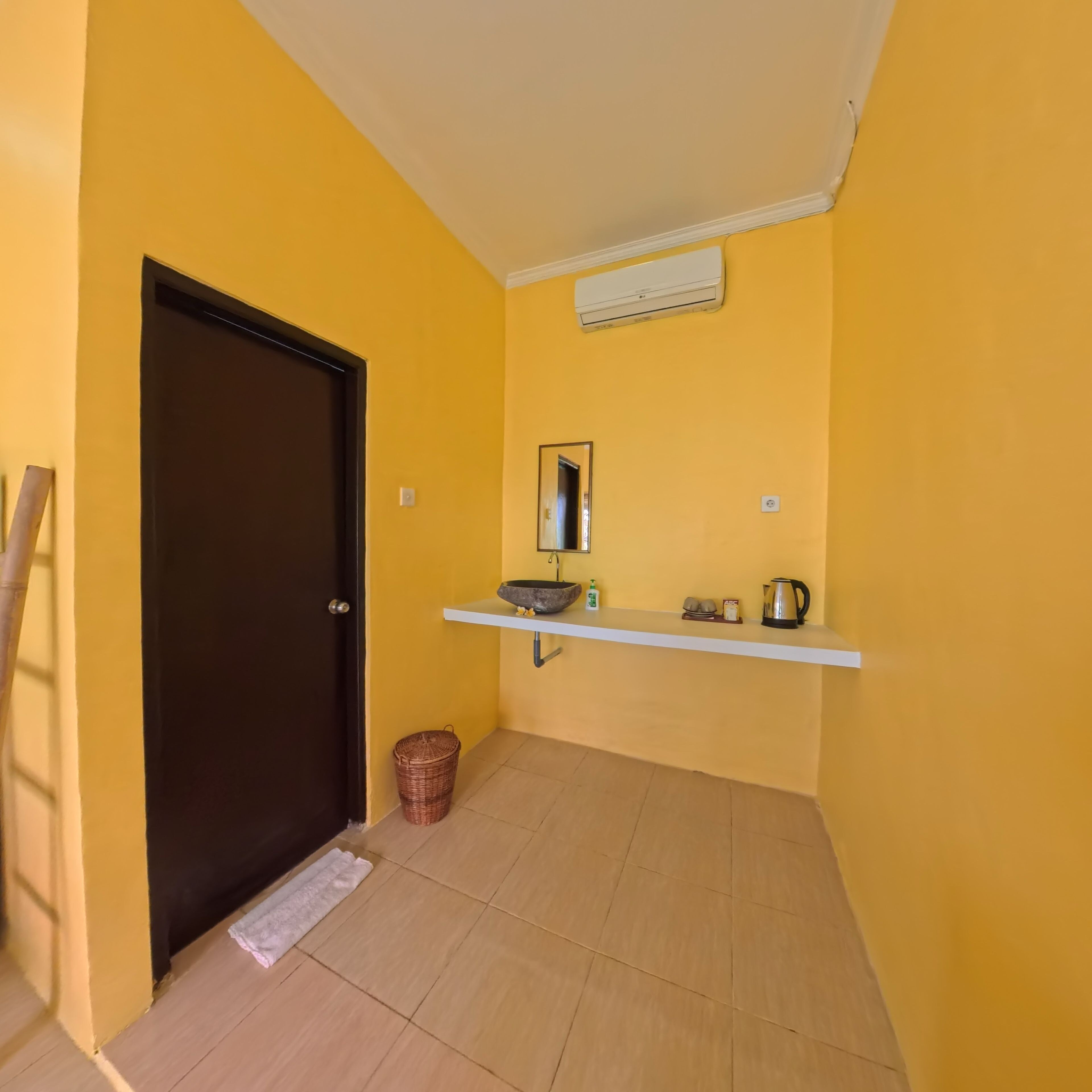 A room with a yellow wall, a dark door, a white shelf with a basin, a kettle, and a trash bin.