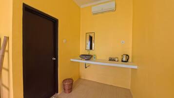 A room with a yellow wall, a dark door, a white shelf with a basin, a kettle, and a trash bin.