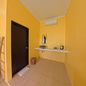 A room with a yellow wall, a dark door, a white shelf with a basin, a kettle, and a trash bin. - Lemon Blossom Guest House (Ubud)