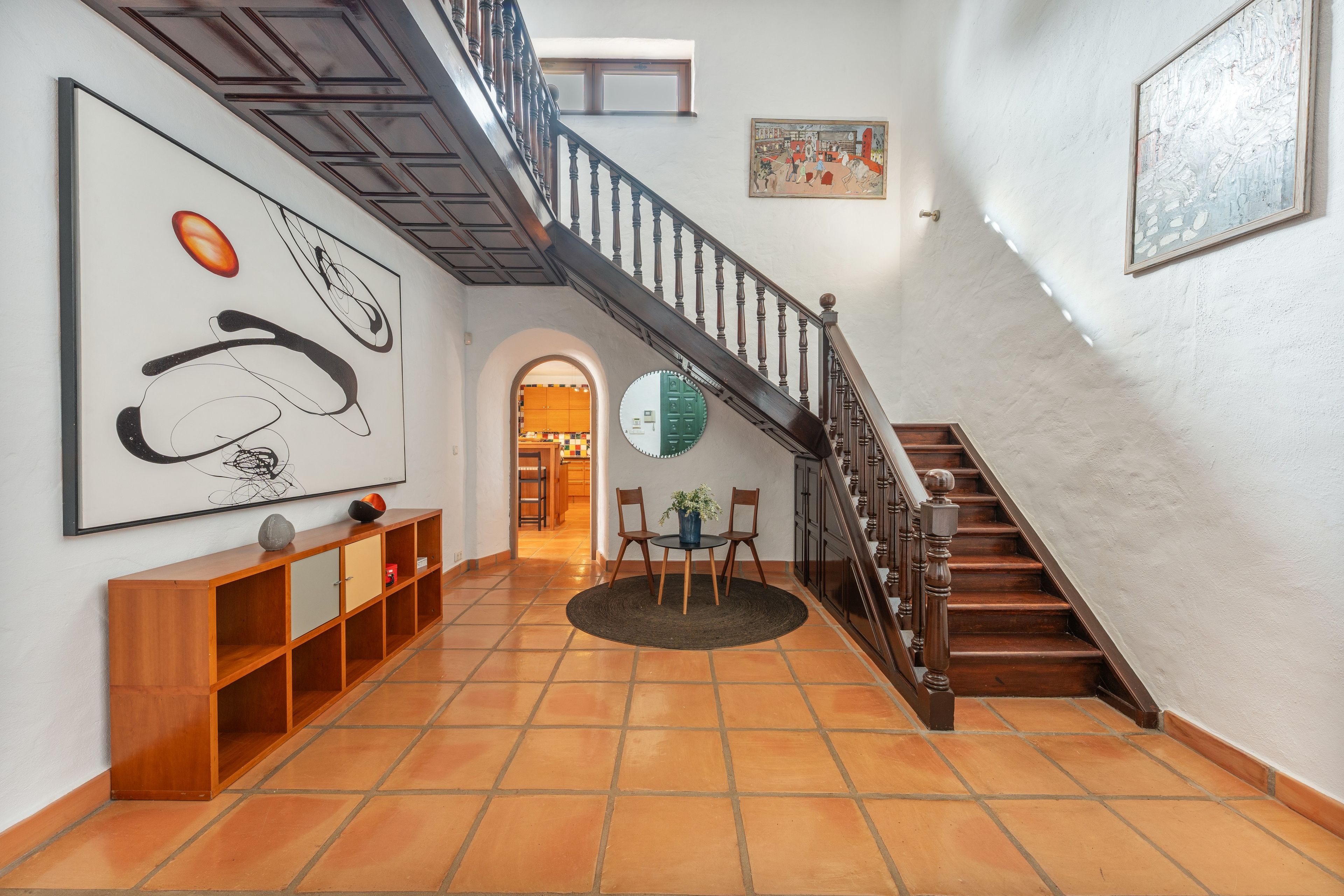 Een gang met een houten trap, een ronde tafel met stoelen en kunstwerken aan de muur.