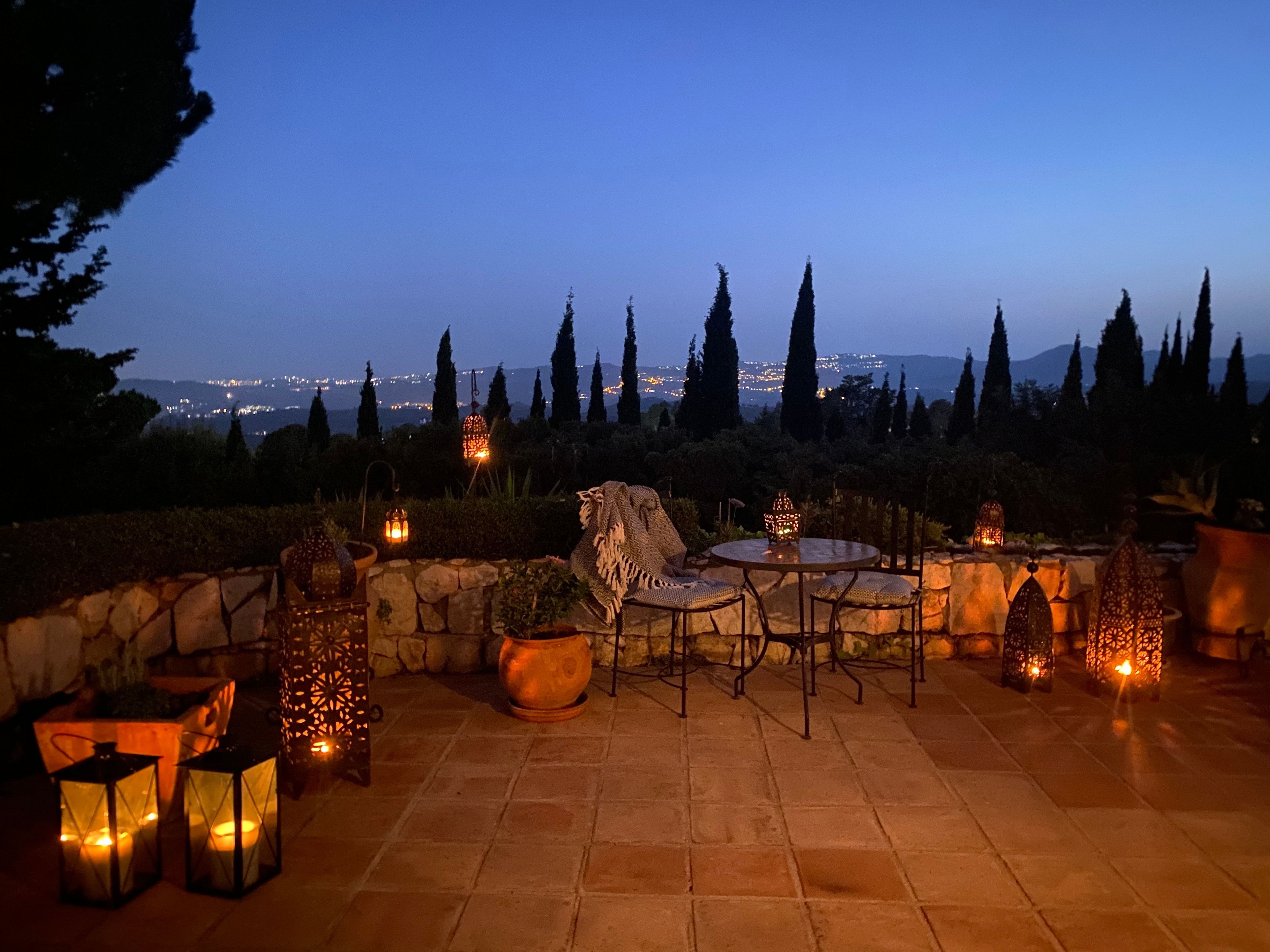 An outdoor patio area with stone flooring, round tables, and chairs, surrounded by decorative lanterns and potted plants.