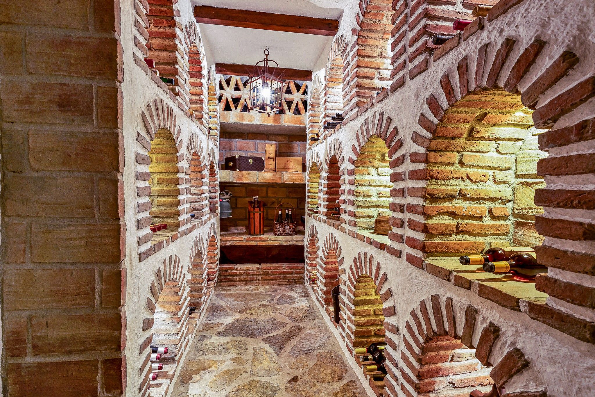 Un estrecho pasaje con paredes de ladrillo y puertas arqueadas, que conduce a una habitación con estantes y botellas.