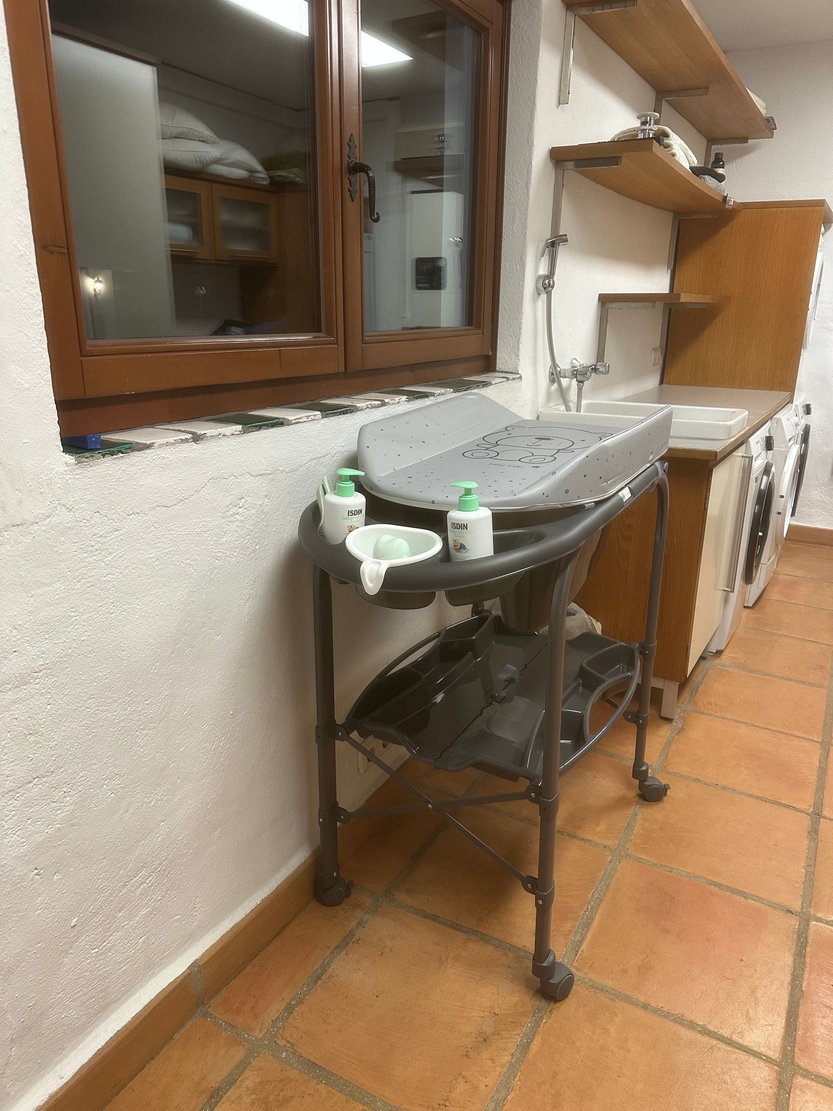 A compact kitchen with a window, a washing machine, and a small table with toiletries.
