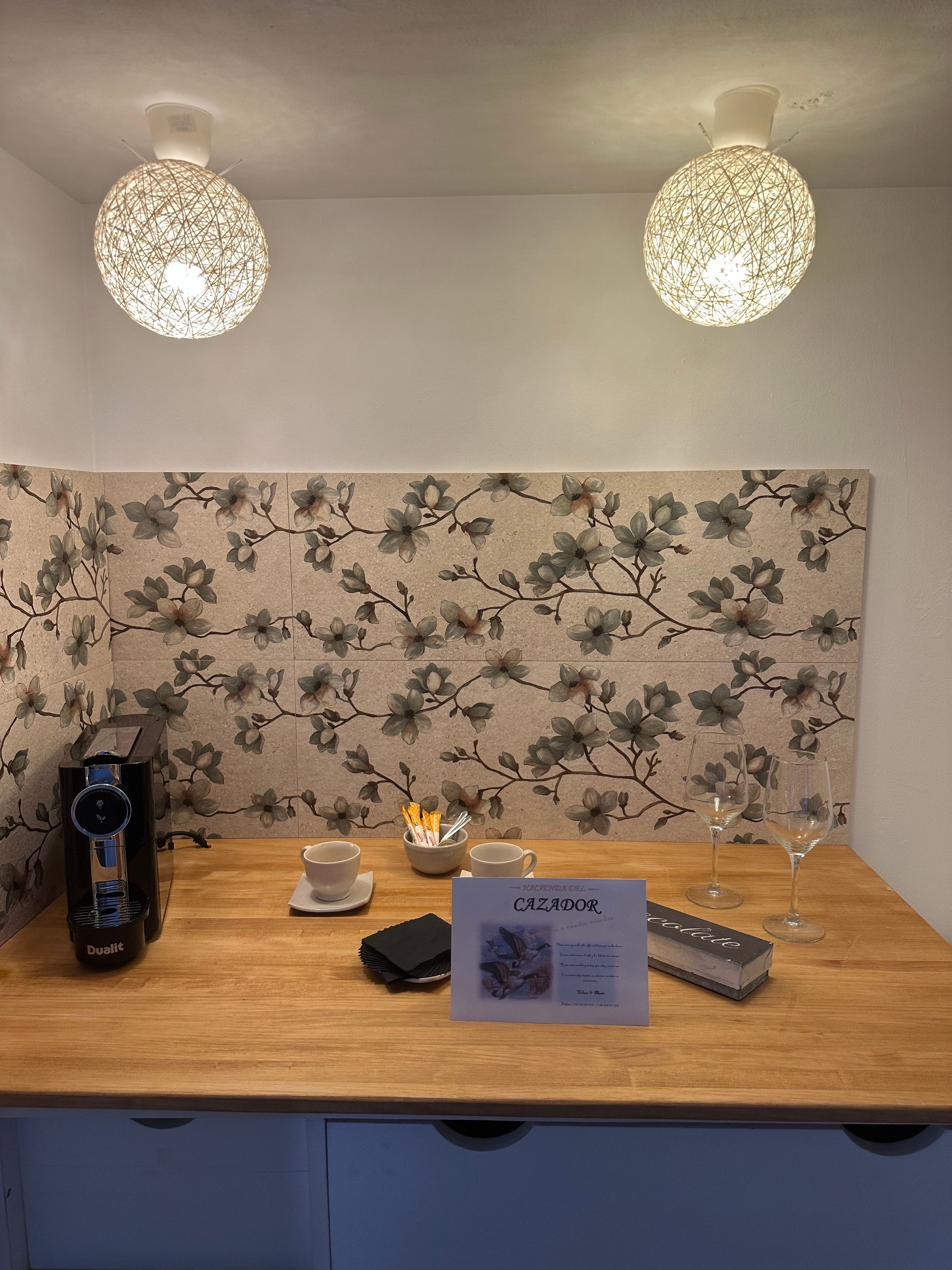 Un mostrador de recepción con un fondo de papel tapiz floral, una máquina de café, dos tazas, una copa y un cartel con el menú.