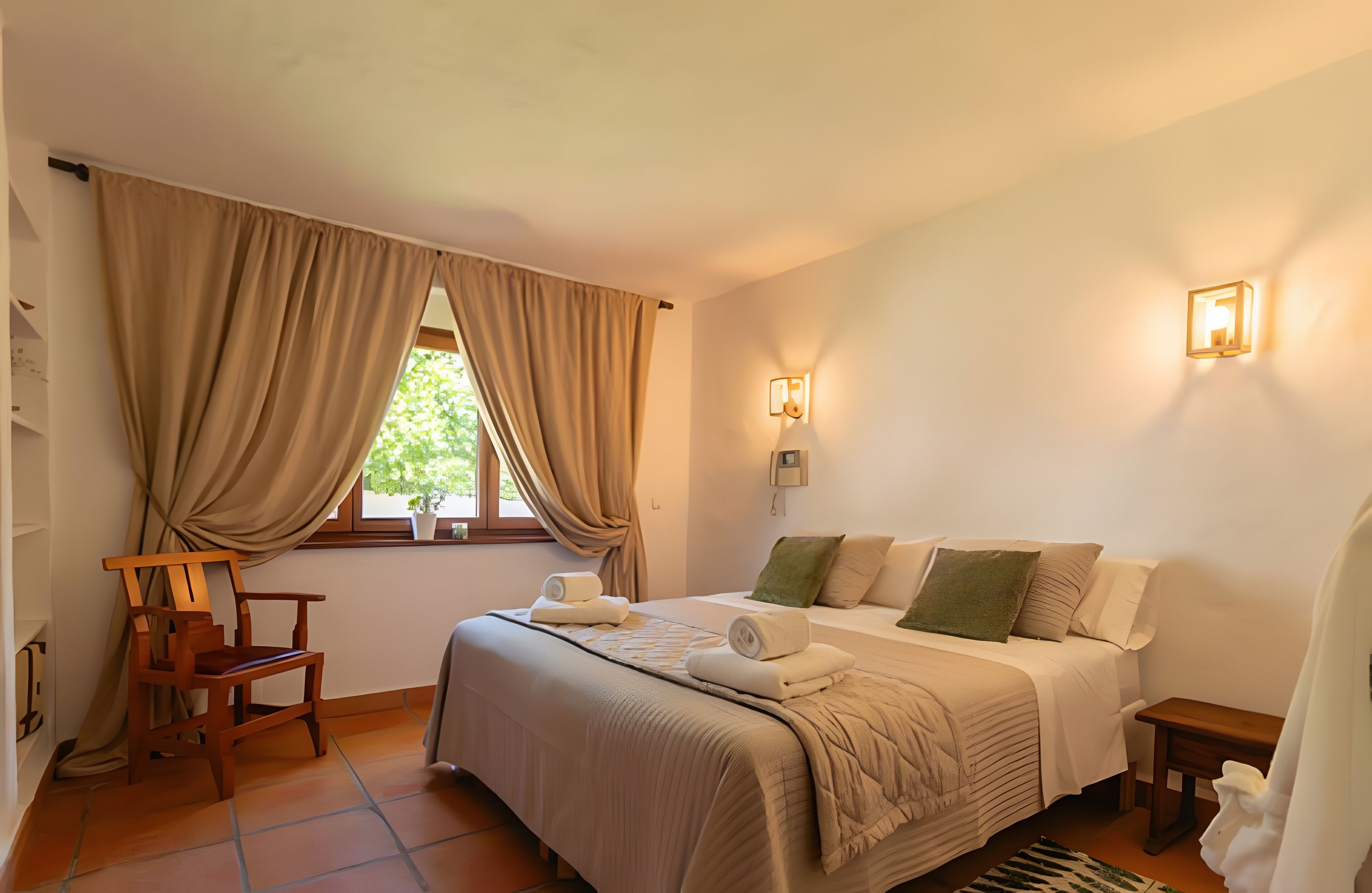 A bedroom with a bed, a chair, a bedside table, a window with curtains, and a wall-mounted light fixture.
