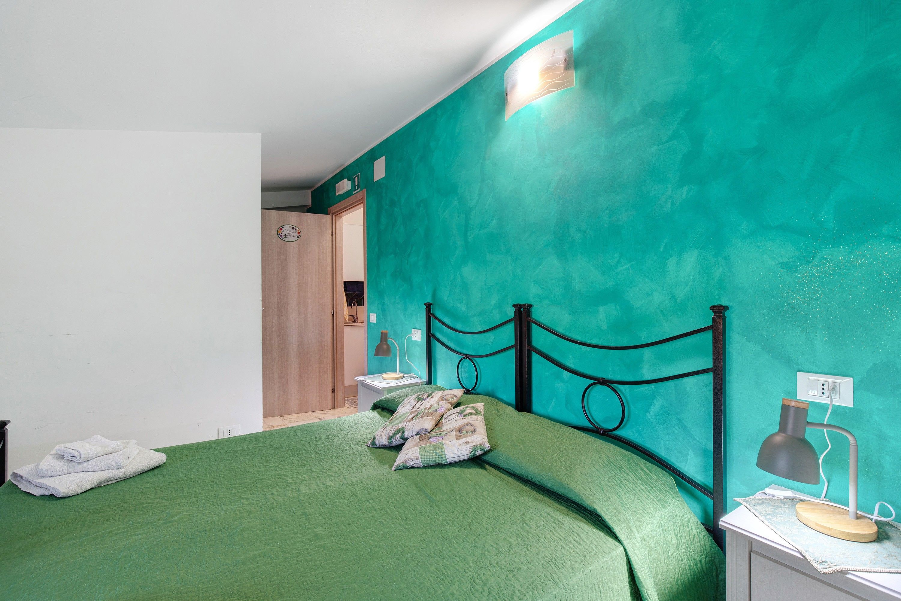 Una habitación con una pared verde azulado, una cama con ropa de cama verde, una mesita de noche con una lámpara y una puerta que da a otra habitación.