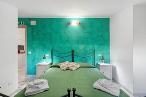 A room with a green textured wall, a bed with a green bedspread, two bedside tables with lamps, and towels on the floor. - Room 'Hope' with Shared Garden, Wi-Fi and Air Conditioning (Roma)