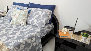 Ein ordentlich bezogenes Bett mit floralem Muster, ein Nachttisch mit einem Buch und einer kleinen Pflanze sowie eine Lampe auf dem Nachtkästchen.