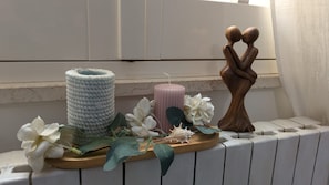 A decorative arrangement on a ledge with a white cylindrical object, a pink candle, white flowers, and a wooden sculpture. - At Giotto, you'll find all the COMFORT YOU NEED (Aradeo)