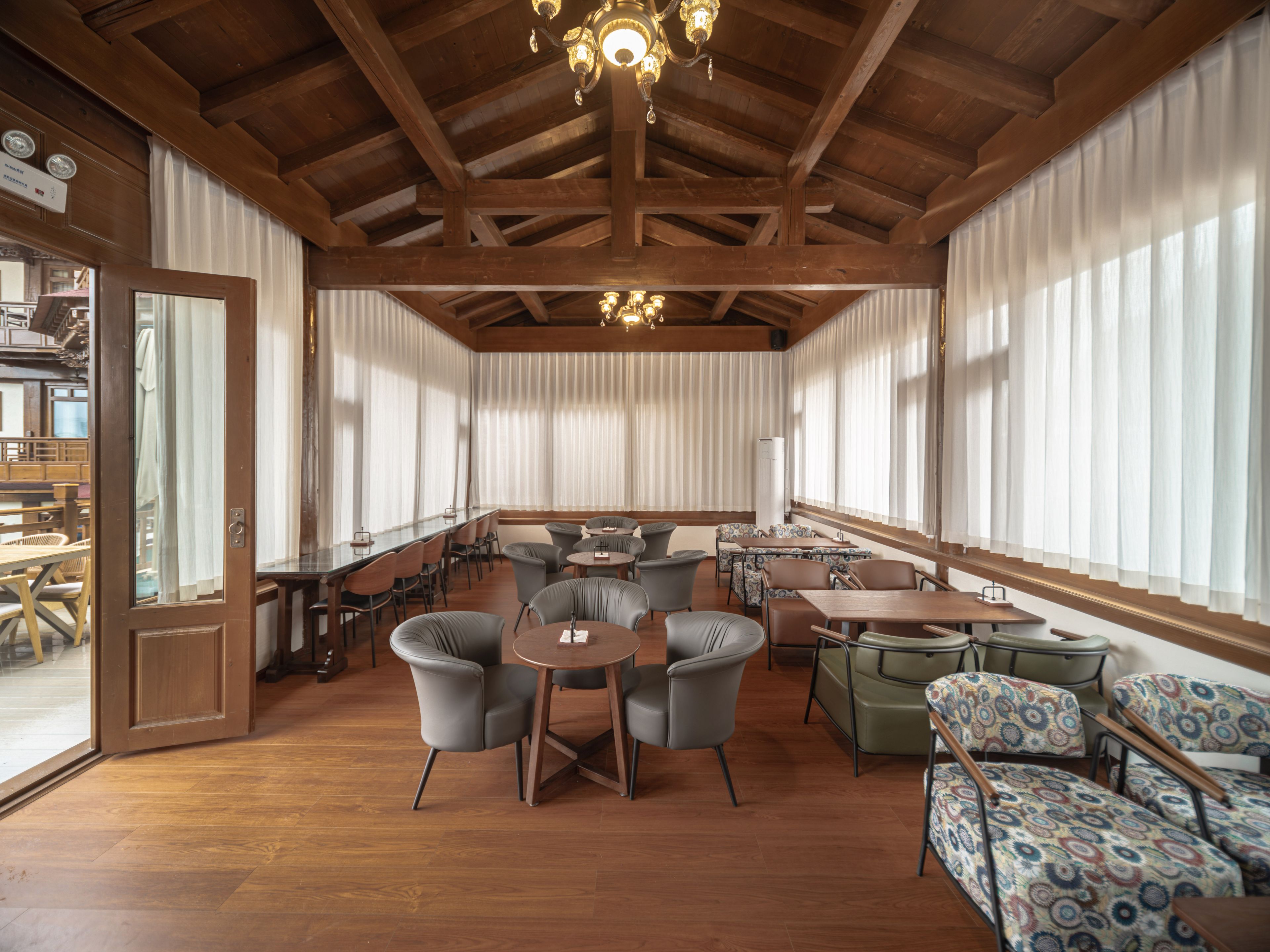 Une pièce spacieuse avec des poutres apparentes, de grandes fenêtres ornées de rideaux légers et divers types de mobilier, notamment des tables et des chaises.