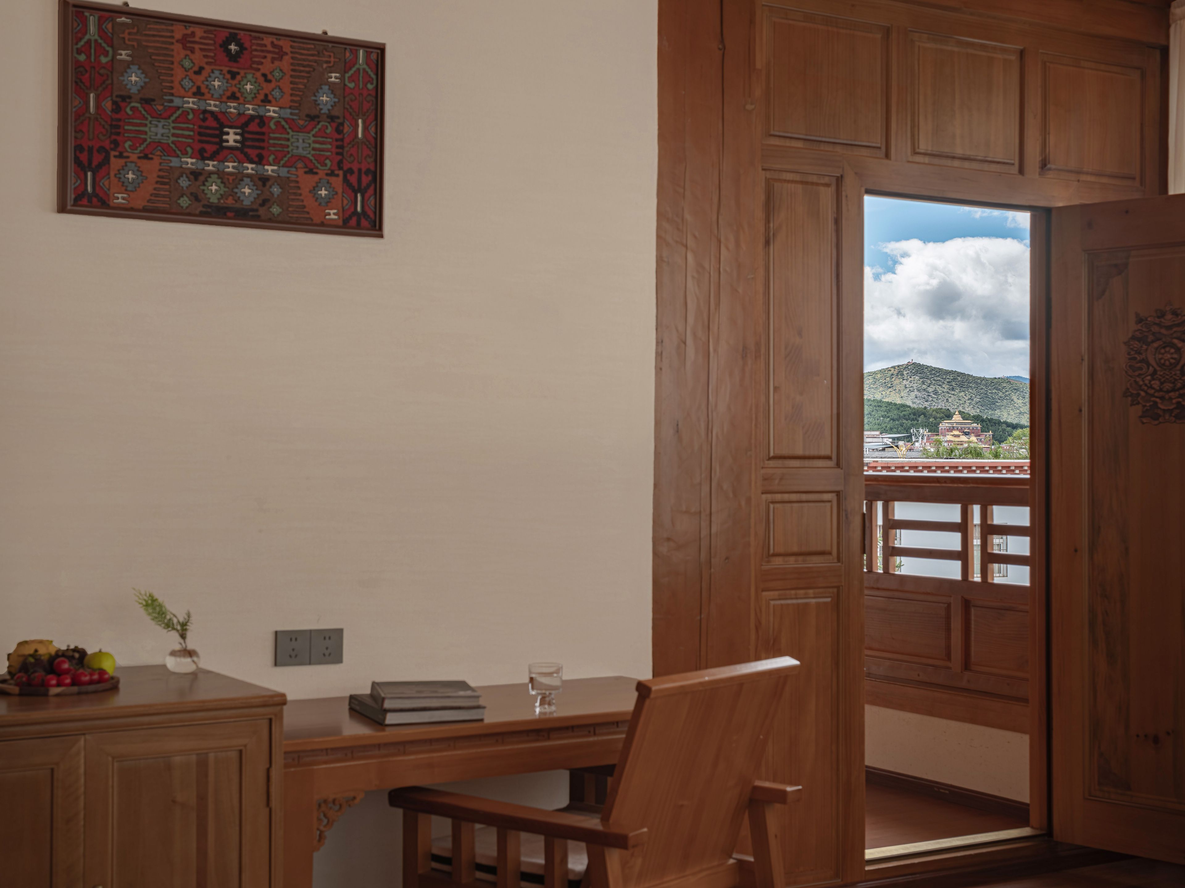 A room with a wooden desk, a chair, a small table with a vase and fruit, and a view of a mountain and temple through a wooden door.