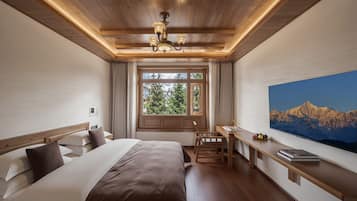 A hotel room with a large bed, a desk, a chair, a window with a view of greenery, and a painting of mountains.