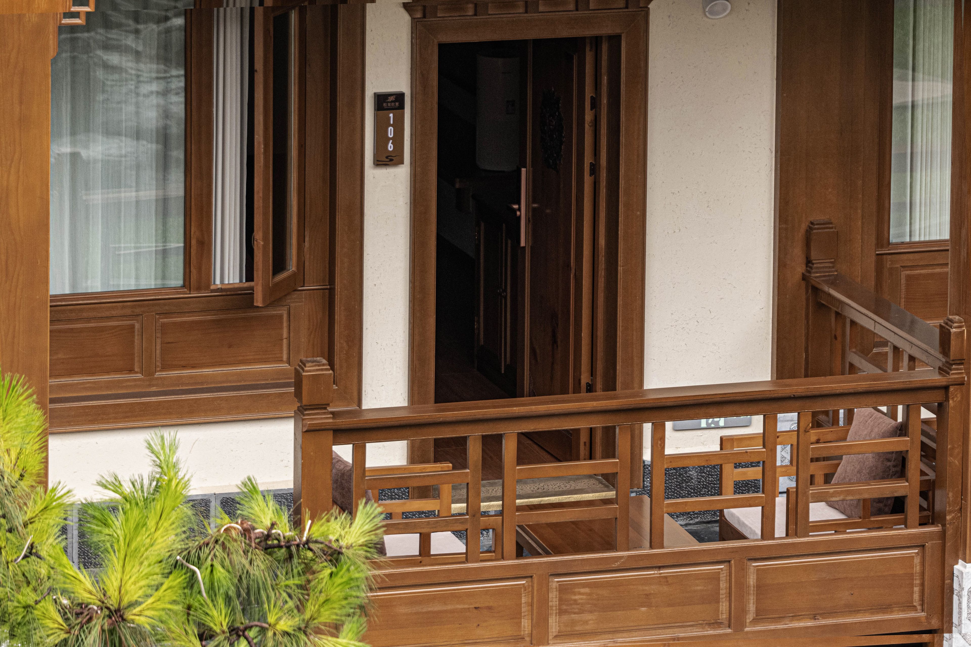 Un balcón de madera con barandilla, una puerta con placa de número y una ventana con cortina.