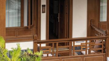 Un balcón de madera con barandilla, una puerta con placa de número y una ventana con cortina.