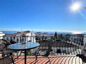 A balcony with a glass table and chairs, overlooking a coastal town with white buildings and a clear blue sky. - Apartment in Nerja With Sea & Mountain Views (Nerja)
